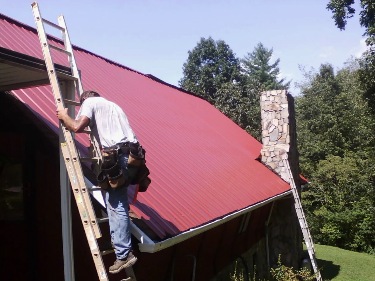 Roofer on a ladder beside a red metal roof and stone chimney — Soffit & Fascia Repair by Roofing Brothers in Park Ridge